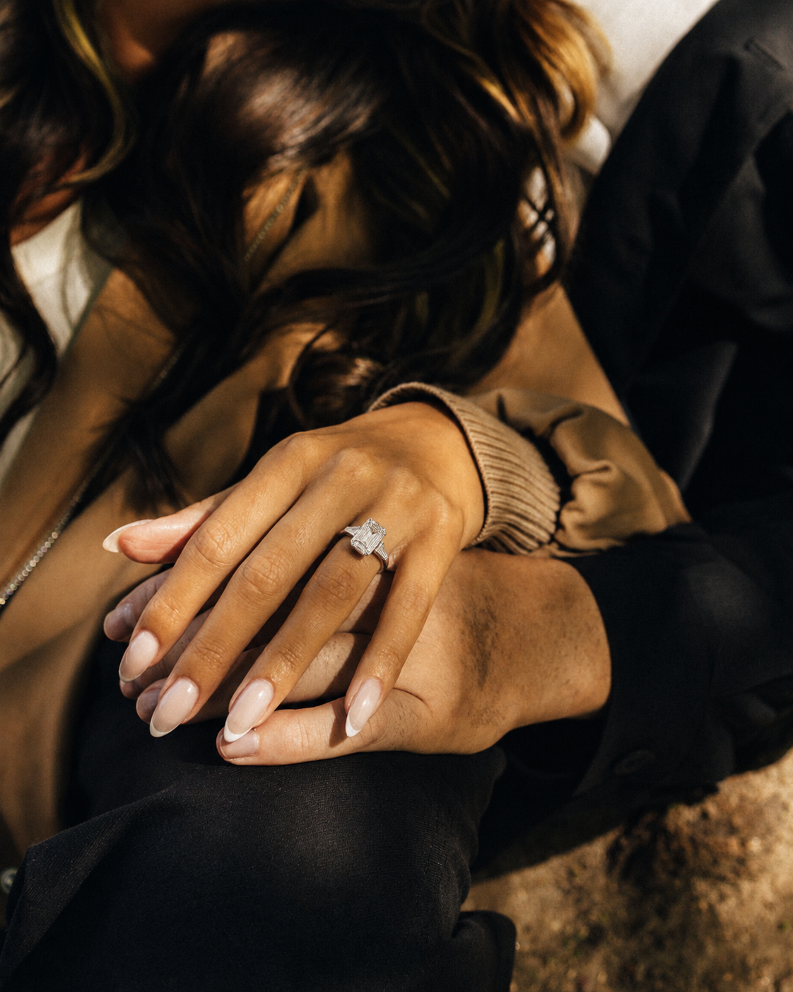 A close-up lifestyle shot of a woman's hand resting on a man's lap, wearing a diamond ring with an emerald-cut center stone.